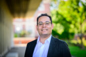 Professor Gabriel Rodriguez wearing a dark suit jacket and shirt standing outside the Education Building on campus.