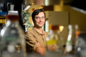 A man in a laboratory, viewed between bottles in the foreground.