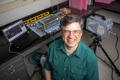 Photo of the researcher in his lab. He is standing in front of a device that sends projectiles into test materials.