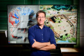 Portrait of Andrew White. He is standing in front of a screen displaying two images: on the left, a LiDAR image of Monks Mound in the ancient city of Cahokia; on the right, an artist's painted interpretation of what the city of Cahokia looked like in its heyday. The painting depicts Cahokia from an aerial perspective.