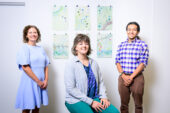 Photo of Sara Benson, e. ainsley and Doxey Kamara in front of a wall with two rows of illustrations behind them.