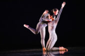Photo of two dancers on stage, dressed in gray unitards with painted feathers on them. A male dancer is kneeling with his arms in the air, and a female dancer is leaning against him with one foot lifted off the ground behind her.