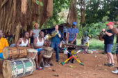 Photo of a group of musicians in a semi-circle outside, most of whom are playing percussion instruments and two of whom are playing French horns.