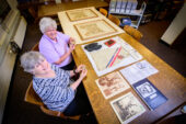 Photo of Susan Jones and Elizabeth Osborn holding gold medals and seated before a table of memorabilia, including track shoes, an Olympic jersey and newspaper clippings and photos.