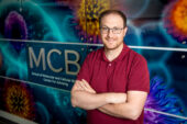 A man stands, arms crossed, in front of a banner with cellular imagery