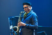 Photo of Charles Lloyd on stage playing a saxophone, wearing a blue jacket and beret and sunglasses.