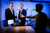 Two researchers stand in suits while a shadowed research subject performs a motor task while wearing a sensor on their hand.