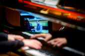 Photo of hands above a piano keyboard with a reflected image of a laptop showing a man seated at a piano.