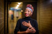 Portrait of Cynthia Oliver wearing a black turtleneck and a colorful headwrap, standing in a hallway with cinder block walls.