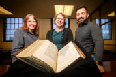 Photo of Cait Coker, Lynne M. Thomas and Elias Petrou standing in front of an open book with Greek text.