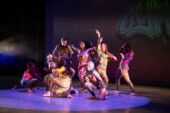 Photo of a group of dancers moving in a spotlight on a stage, wearing multicolored leotards.