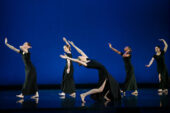 Photo of dancers in black dresses with one in the foreground leaning back with her torso and arms, and four others in the background facing the opposite direction.