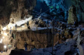 Image of the interior of the cave and the massive trench with people standing at different levels and looking into the trench. The cave is dark and you can see the grid of guidelines used to plot the location of items found in the dig. There are bright worklights overhead.