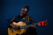 Photo of Okaidja Afroso holding his guitar and singing with his eyes closed.
