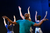 Photo of a Black man with a graying goatee dancing with outstretched arms, while several female dancers with raised arms dance in the background.