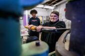 Photo of a woman pulling a lever on a letterpress while another student watches. They are framed by parts of the machine in the foreground.