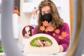 Photo of Tamar Dallal wearing a face mask and seated at a table, with a low, curving vase in the foreground and twig of red maple leaves coming from the vase.