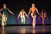 Photo of dancers on a stage, two seen from the front and two from the back, wearing khaki pants and t-shirts or tank tops. They have both feet off the ground and their arms outstretched along their sides.