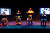 Photo of four actors on stage in front of music stands, three men and one woman. The two on the ends seated and the two in the middle standing.There are two video boards with abstract images behind them.