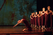 Photo of dancers lined up on stage in matching rust-colored jumpsuits, with one dancer kneeling and striking a pose to the side.