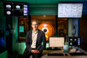 Researcher sits on a desk with readouts on computer monitors surrounding him and a magnetic resonance imaging device in the background.