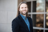 U. of I. graduate student Nathan Tanner standing outdoors next to a building's windows on the university campus