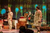 A white man in a gold crown, sash and filthy white pants and a black man in a furry coat stare at each other on stage in front of an armchair and piles of garbage.