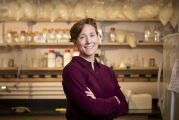 Kate Clancy stands in her laboratory. She is wearing a maroon shirt, has her arms crossed, and she is smiling.