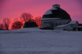 Red and purple sky behind a barn with snow-covered fields in the foreground.