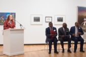 Krannert Art Museum Director Kathleen Harleman stands at a podium as, Kenyan Ambassador Robinson Njeru Githae, U. of I. Chancellor Robert Jones, Director General of the National Museums of Kenya Mzalendo Kibunjia sit next to each other in chairs at Krannert Art Museum, University of Illinois at Urbana-Champaign.