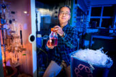 Photo of the researcher in his laboratory with starting materials and holding a glass jar full of the end product, ethylbenzene.