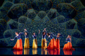 Photo of seven dancers onstage wearing blue tops and orange or yellow flowing skirts. The backdrop is a Persian design.