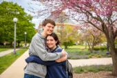 Male and female student embracing on the quad with flowering redbud tree and the ACES library in the background. Photo by Michelle Hassel
