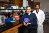 Two men in a lab. The seated man holds a hologram projection of a brain.