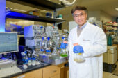 Portrait of Yong-Su Jin in the lab wearing a white lab coat and holding two flasks.