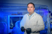 A man in a lab coat and gloves holds a culture dish next to a fume hood in a lab.