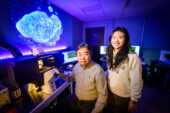 Researchers positioned behind a microscope used in the research and in from of a projected image of model cancer cell.