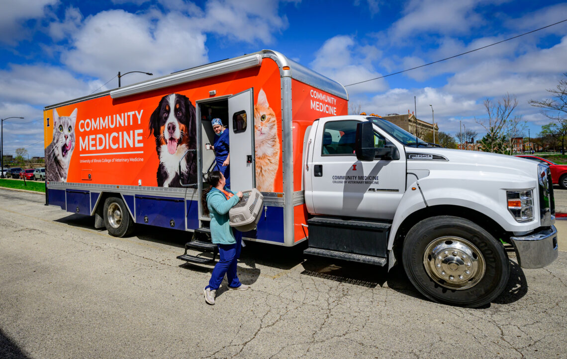 Photo outside the mobile veterinary clinic, a long orange truck with the words "Community Medicine" and large photos of dogs and cats emblazoned on the side. 