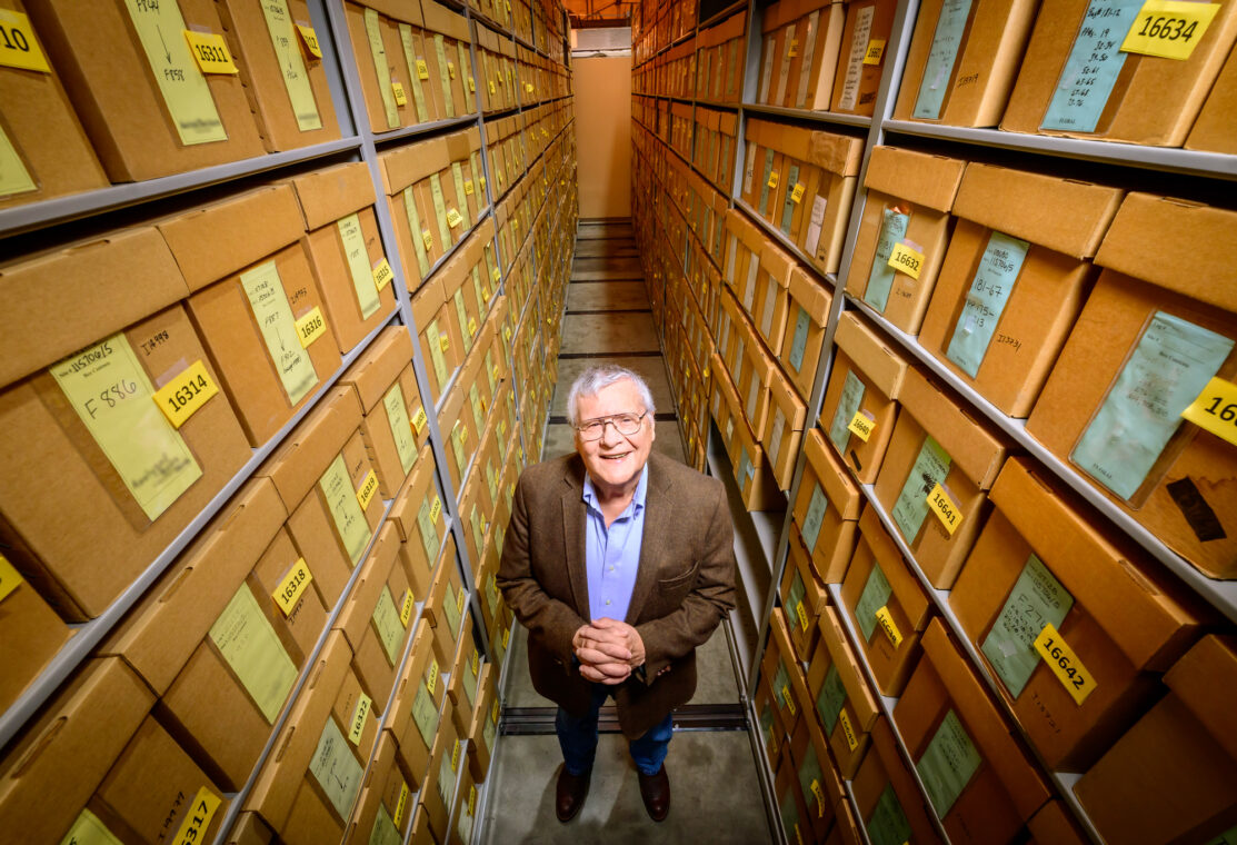 Photo of Thomas Emerson inside a storage room. Two walls of storage boxes extend far above his head. He is clasping his hands together and looking up at the photographer.