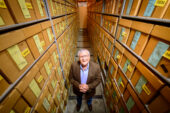Photo of Thomas Emerson inside a storage room. Two walls of storage boxes extend far above his head. He is clasping his hands together and looking up at the photographer.