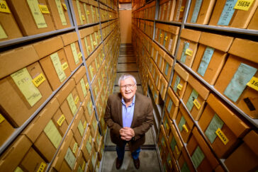 Photo of Thomas Emerson inside a storage room. Two walls of storage boxes extend far above his head. He is clasping his hands together and looking up at the photographer.