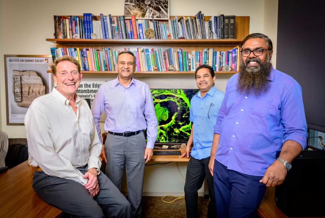 Four men stand in front of a bookshelf and a screen with a microscope image displayed on it