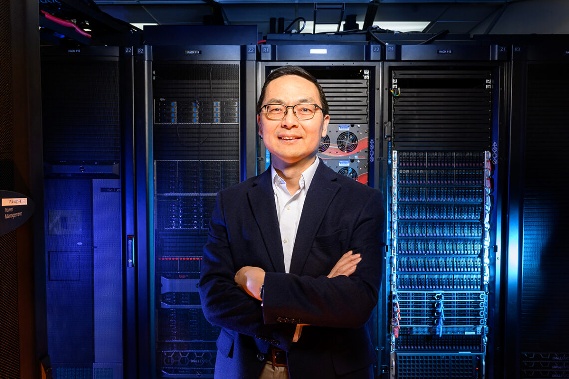 A man in a suit stands in front of a room full of computer servers, lit by blue light