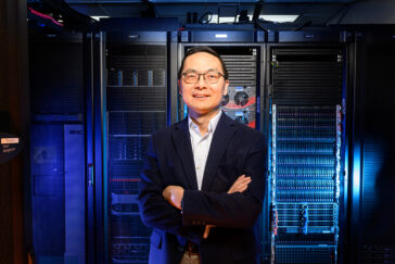A man in a suit stands in front of a room full of computer servers, lit by blue light