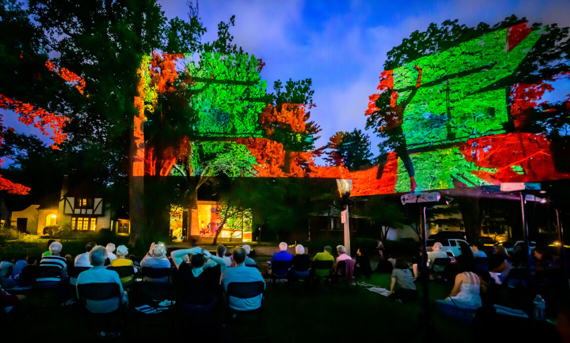 Photo of people seated in chairs looking up at a colorful projection onto the side of a house and trees.