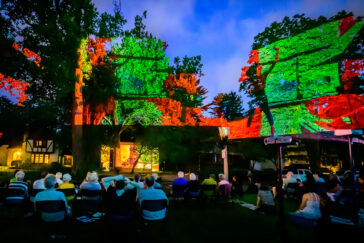 Photo of people seated in chairs looking up at a colorful projection onto the side of a house and trees.