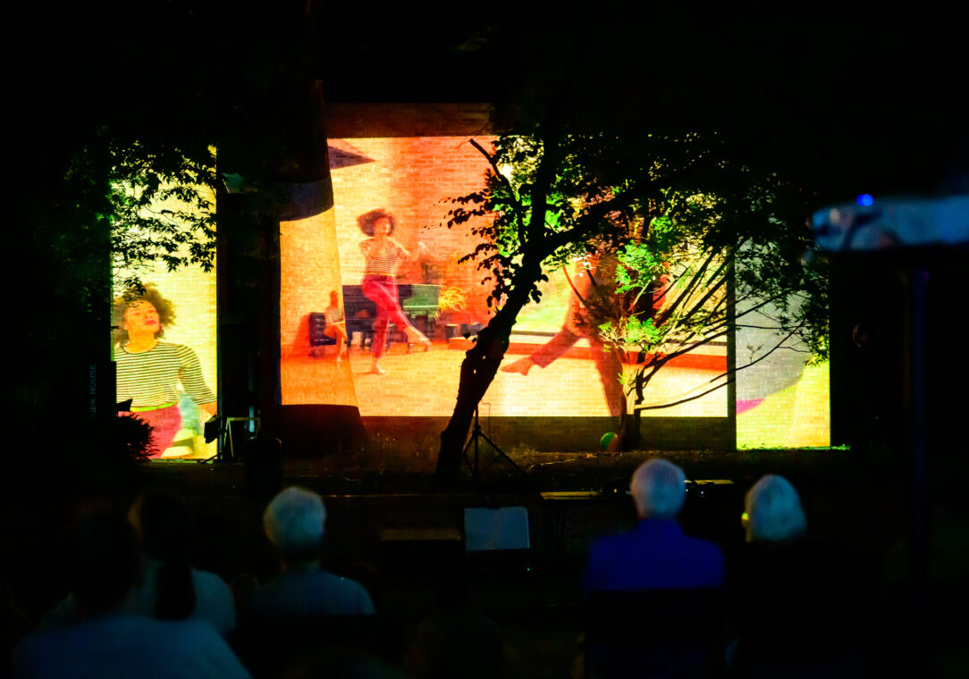 Photo of a video of a dancer projected onto the exterior of a house, with a tree in the middle of one of the projections.