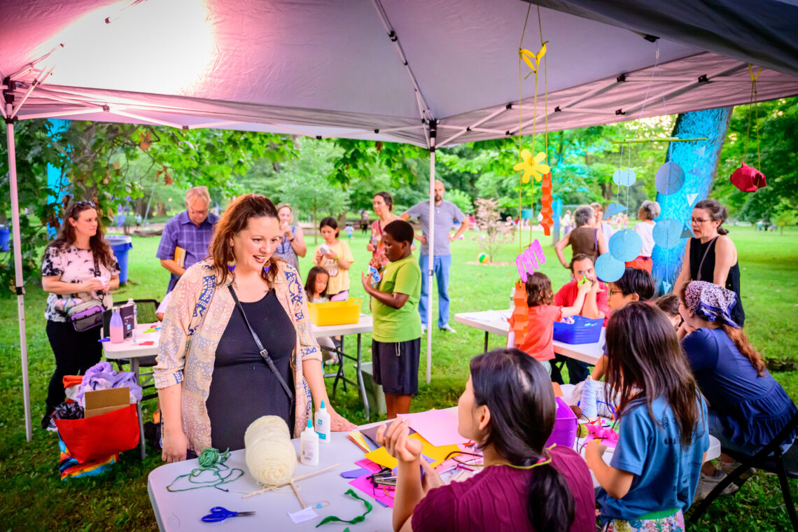 Photo of a woman standing in front of a craft table underneath a tent in a park, while other people in the background are busy with other crafts.