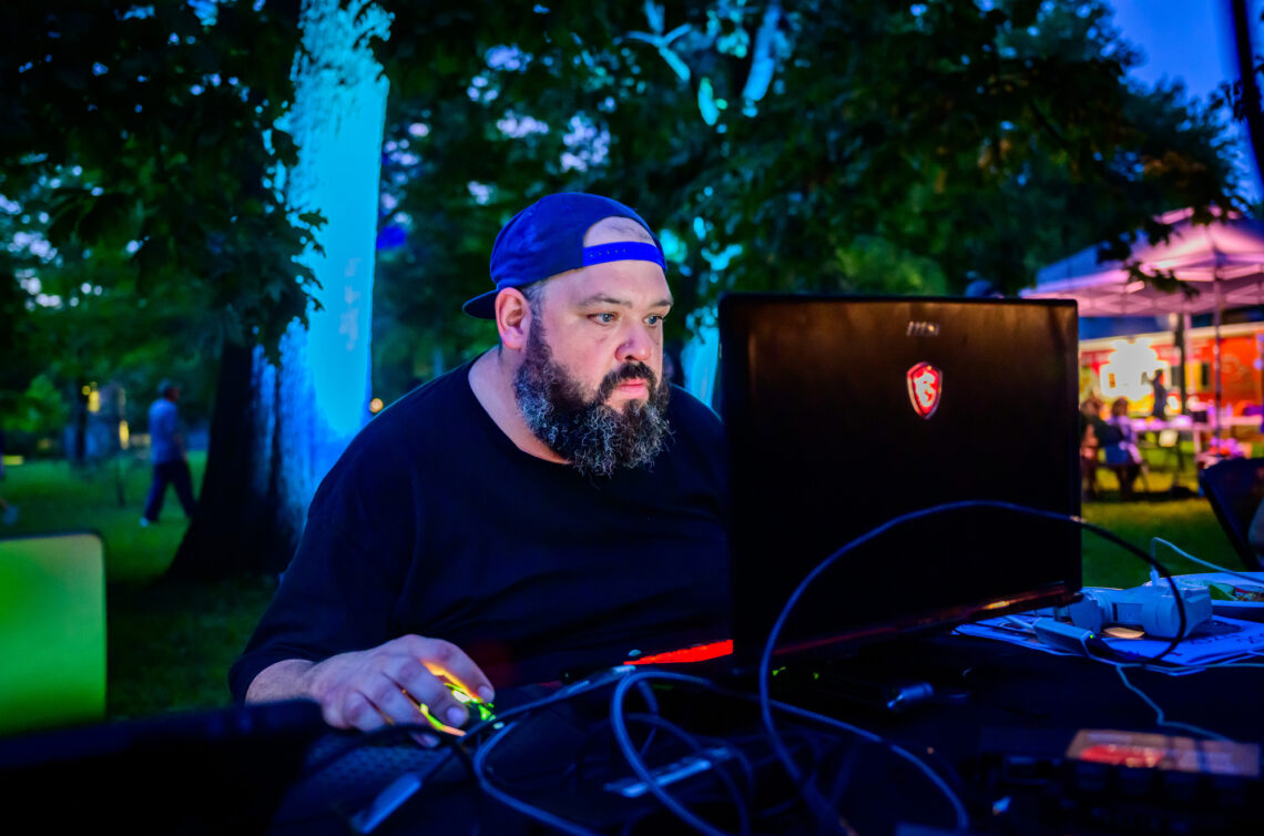 Photo of a bearded man wearing a backwards baseball cap sitting before a computer outside at dusk.