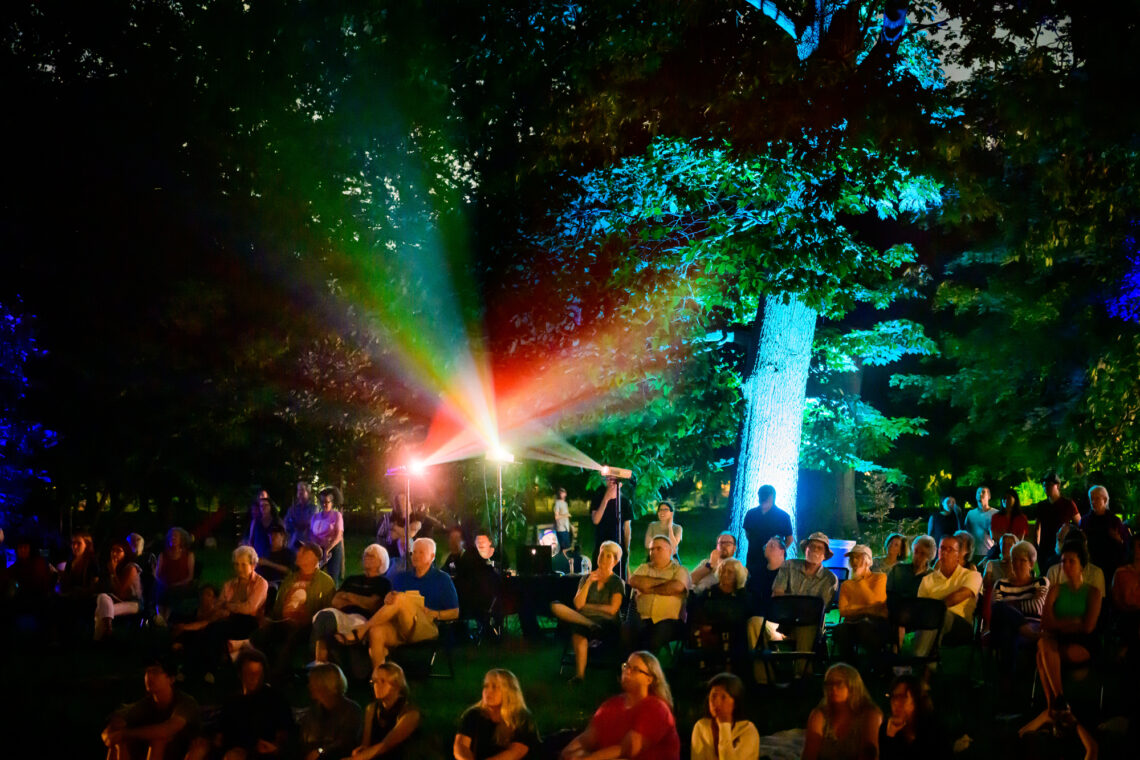 Photo of people seated in a park at dusk with projectors beaming light behind them.